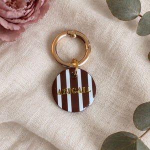 May include: A circular keychain featuring a brown and white vertical striped pattern with the name ABIGAIL embossed in gold lettering. The tag is attached to a polished gold-toned metal ring clasp, providing a secure attachment point.