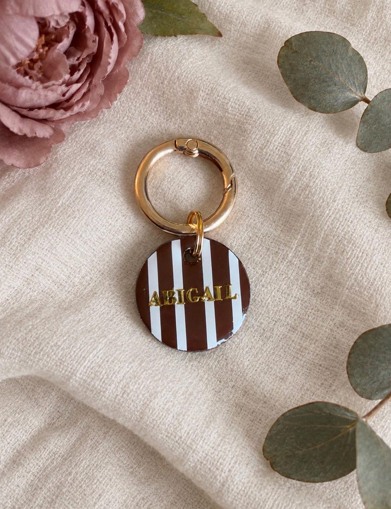 May include: A circular keychain featuring a brown and white vertical striped pattern with the name ABIGAIL embossed in gold lettering. The tag is attached to a polished gold-toned metal ring clasp, providing a secure attachment point.