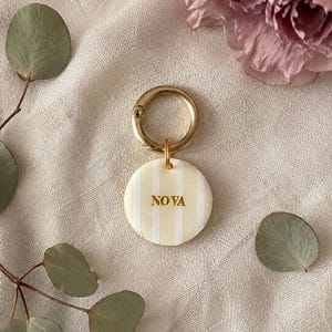 May include: A circular keychain featuring a cream and white vertical striped pattern with the name NOVA embossed in gold lettering. The disc is attached to a polished gold-toned metal ring clasp, providing a secure attachment point for keys.