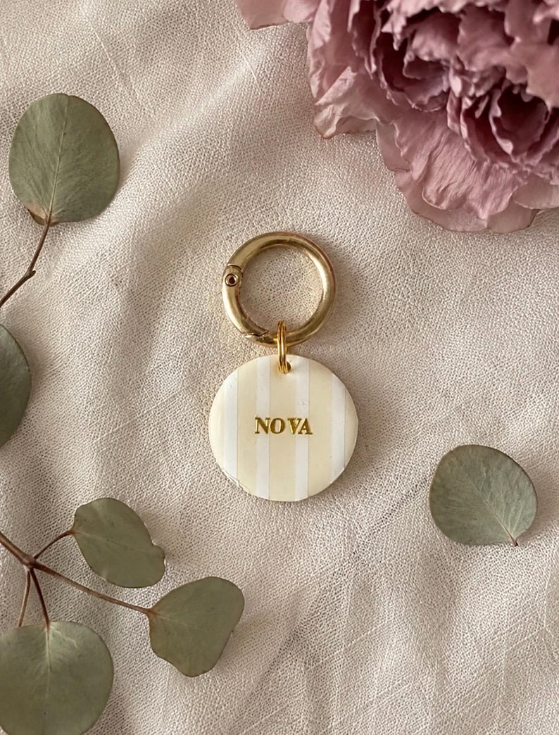 May include: A circular keychain featuring a cream and white vertical striped pattern with the name NOVA embossed in gold lettering. The disc is attached to a polished gold-toned metal ring clasp, providing a secure attachment point for keys.
