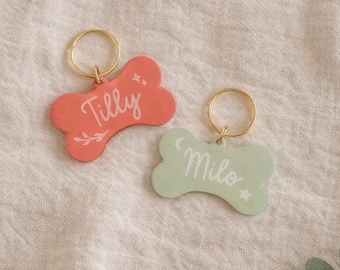 May include: Two bone-shaped pet tags with gold-tone metal rings. One tag is coral-colored with the white script name Tilly, a small leaf, and star icons. The other tag is sage green with the white script name Milo, a crescent moon, and a star.