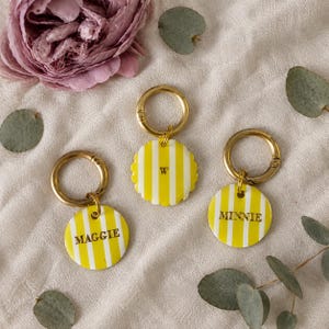 May include: Three circular keychains with gold-toned metal rings, each featuring a yellow and white vertical striped pattern. One keychain is engraved with the name MAGGIE, one with the name MINNIE, and one with the single letter W.