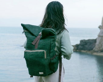 May include: A person wearing a green and brown backpack stands on a cliff overlooking a body of water. The person is wearing a light green shirt and white shorts. The backpack has a flap that folds over the top.
