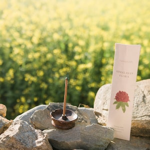 May include: A lit incense stick in a bronze holder, with a package of "Himalayan Flora" incense. The package features a red flower illustration. The scene is set outdoors on a stone wall with a blurred yellow flower background.