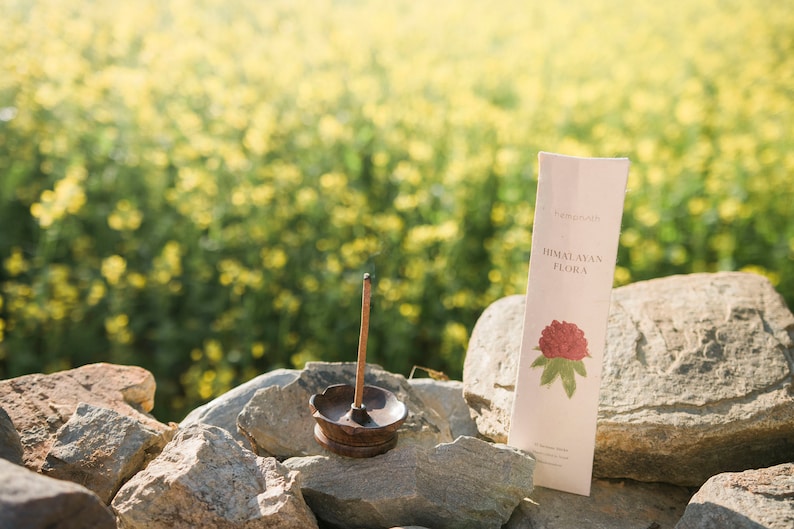 May include: A lit incense stick in a bronze holder, with a package of "Himalayan Flora" incense. The package features a red flower illustration. The scene is set outdoors on a stone wall with a blurred yellow flower background.