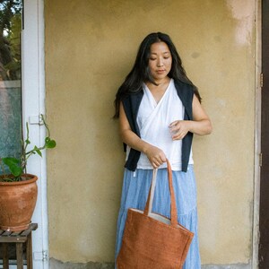 May include: A rectangular, rust-colored tote bag with two matching fabric handles. The bag features a textured surface and a small white rectangular label stitched near the bottom corner. The bag is held by its handles, displaying its vertical orientation.