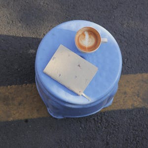 A handmade paper notebook placed on a small blue table next to a coffee cup, capturing a moment of calm creativity. This image evokes the use of the notebook as a daily journal, mindful writing notebook, or eco friendly sketchbook