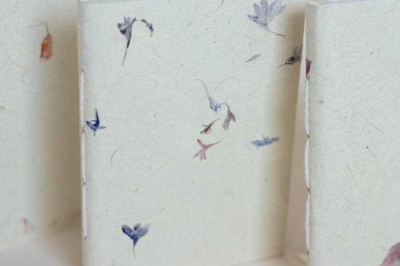A close, detailed photo of notebook covers with small pressed petals embedded into handmade paper. Perfect for showing the unique texture of artisan paper notebooks, botanical stationery, and eco conscious journals.