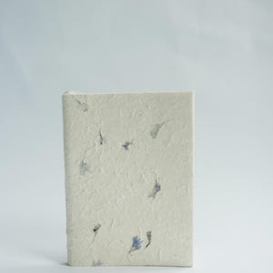 A single recycled paper notebook standing upright with visible flower petals embedded in the cover. The clean background draws attention to the handmade quality associated with lokta paper journals and sustainable stationery.