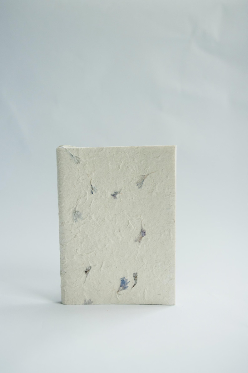 A single recycled paper notebook standing upright with visible flower petals embedded in the cover. The clean background draws attention to the handmade quality associated with lokta paper journals and sustainable stationery.