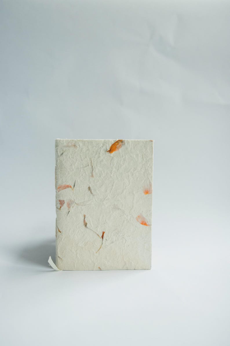 A minimal image showing a handmade lokta paper notebook standing upright against a clean white backdrop. The soft pressed petals and textured cover reflect the feel of a sustainable journal, recycled paper notebook, and handcrafted writing book.