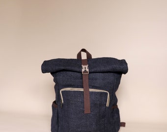 May include: A dark blue canvas backpack with brown straps and a roll-top closure. The backpack has a zippered pocket on the front and a small label on the bottom.