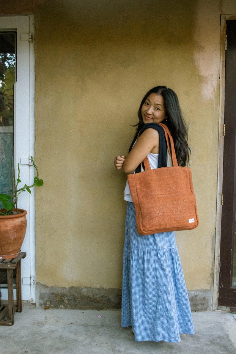 May include: A rectangular, burnt-orange tote bag with two shoulder straps and a small white rectangular label on the lower right corner. The bag features a textured, woven fabric construction and is carried over the shoulder.