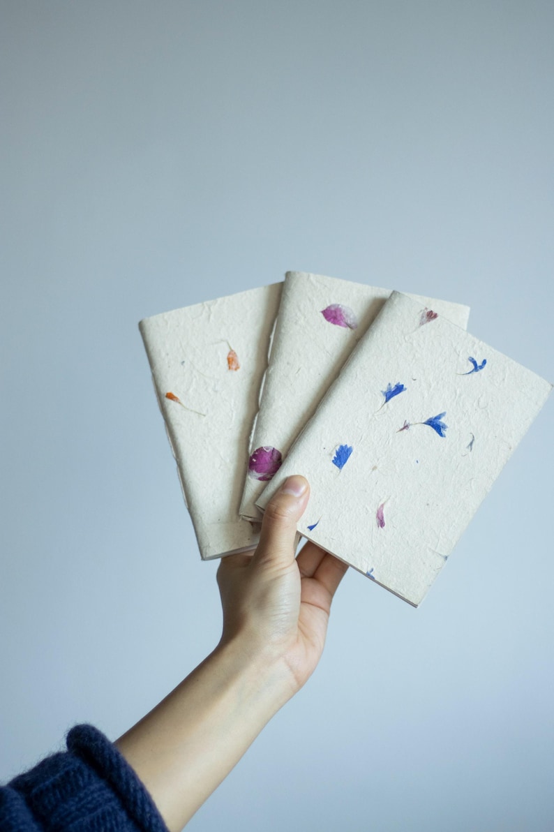 A person holding three handmade paper notebooks in their hand, showing the natural texture and pressed petals of the lokta paper covers. The notebooks have soft, organic edges and subtle botanical inclusions, highlighting their nature-inspired design. This image expresses the feel of an eco friendly notebook, artisan journal, and sustainable stationery piece made from recycled handmade paper.