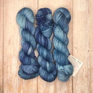 Op de afbeelding: Drie strengen handgeverfd garen in blauwtinten, van licht turquoise tot diep indigo. Het garen ligt op een licht houten oppervlak. Een klein label met de tekst "Blue Ocean" is bevestigd.