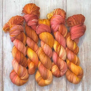 Op de afbeelding: Close-up van verschillende strengen handgeverfd garen in tinten oranje, geel en roze. Het garen is gedraaid in een touwachtige vorm, waarbij de kleuren in elkaar overlopen. Het garen is gerangschikt op een lichtgekleurd houten oppervlak.