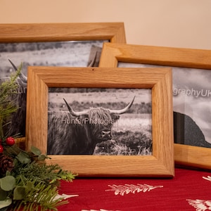 May include: Three wooden picture frames with black and white photographs of a highland cow and a mountain landscape. The frames are on a red tablecloth with a white pattern.