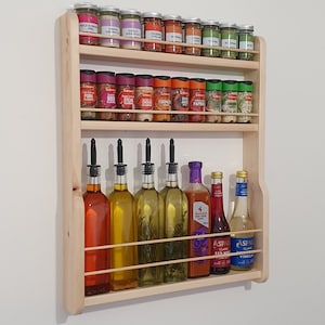 May include: A wooden spice rack with three shelves, filled with various spices and bottles of oil and vinegar. The rack is mounted on a white wall.