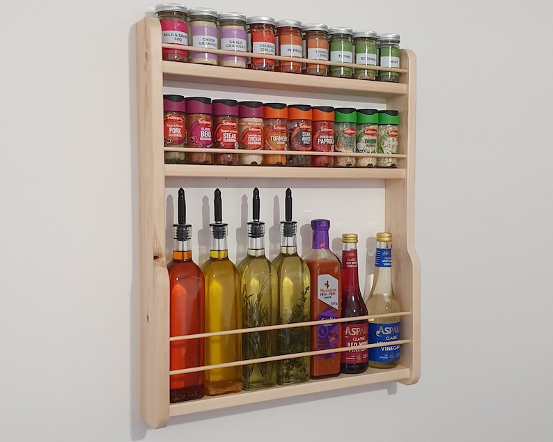 May include: A wooden spice rack with three shelves, filled with various spices and bottles of oil and vinegar. The rack is mounted on a white wall.