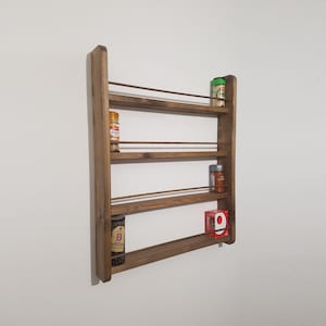 May include: A wooden spice rack with four shelves and copper rails. The rack is mounted on a white wall and holds various spice jars and a red box.