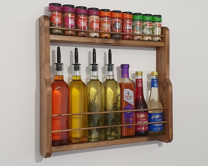 May include: A wooden wall-mounted spice rack with two shelves. The top shelf holds various spice jars with labels like "Pork", "Steak", "Chicken", "Turmeric", "Paprika", and "Parsley". The bottom shelf holds bottles of oil, vinegar, and red wine vinegar. The bottles have labels like "Aspall Classic Red Wine Vinegar" and "Aspall Classic Vinegar".