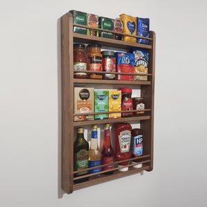 May include: A wall-mounted wooden spice rack with three horizontal shelves, each featuring a thin wooden safety rail. The rack is filled with various food items, including boxes of tea, jars of sauces, bottles of oil, and condiments like Heinz ketchup.