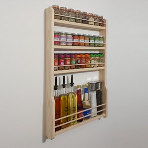 May include: A wooden spice rack with four shelves, holding various glass jars of spices and bottles of oil and vinegar. The rack is mounted on a white wall.