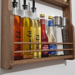 May include: Wooden wall-mounted shelf with six glass bottles and two glass shakers. The bottles contain various liquids, including chili oil, garlic oil, olive oil, red wine vinegar, and white wine vinegar. The shakers contain salt and pepper.