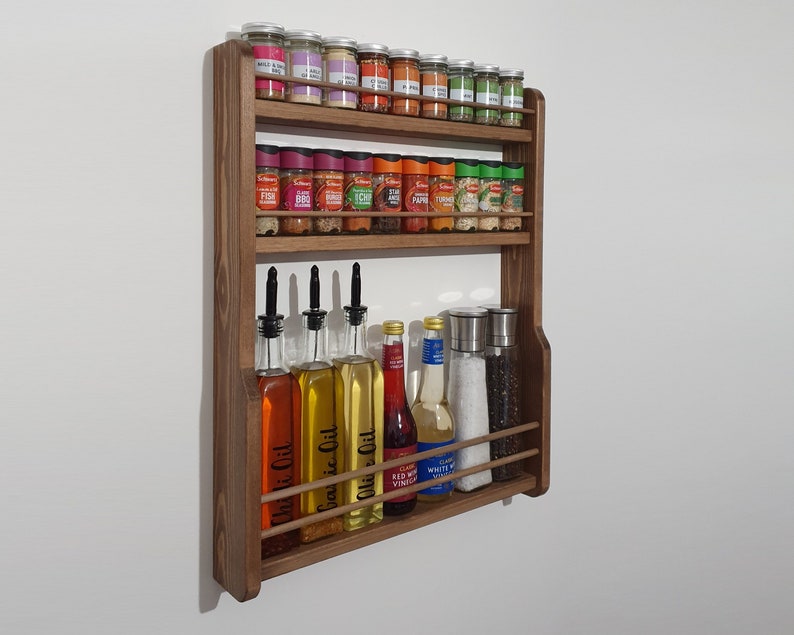 May include: A wooden spice rack with two shelves and a bottom rack for bottles. The top shelf is filled with small glass jars of spices, while the bottom shelf holds bottles of oil and vinegar. The rack is mounted on a white wall.