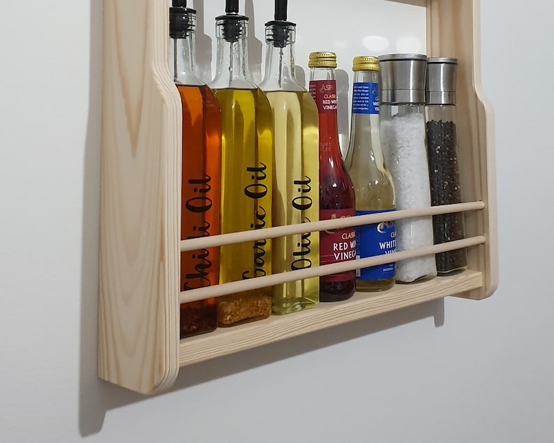 May include: A wooden wall-mounted shelf with six glass bottles and two glass jars. The bottles contain various oils, including chili oil, garlic oil, and olive oil. The jars contain white salt and black peppercorns. The labels on the bottles are black and white with white text.