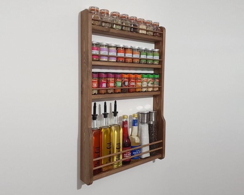 May include: A wooden spice rack with four shelves, holding various glass jars of spices and bottles of oil and vinegar. The rack is mounted on a white wall.