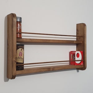 May include: A brown wooden wall-mounted spice rack with two shelves and metal rails. The rack is holding two spice jars and a box of spices. The jars are labeled "Tesco Cayenne" and "Tesco Black Peppercorns". The box is labeled "Sainsbury's Garlic & Herb".