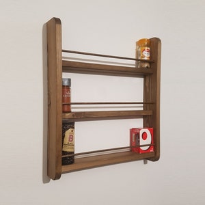 May include: A wooden spice rack with three shelves. The rack is mounted on a white wall. There are three glass jars of spices on the shelves, and a red box of crackers on the bottom shelf.