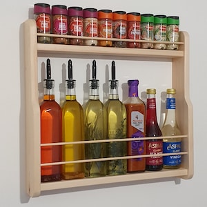 May include: A wooden wall-mounted spice rack with two shelves. The top shelf holds various spice jars with labels like "Pork Steak", "Chicken", "Turmeric", "Paprika", and "Garlic". The bottom shelf holds bottles of oil and vinegar.