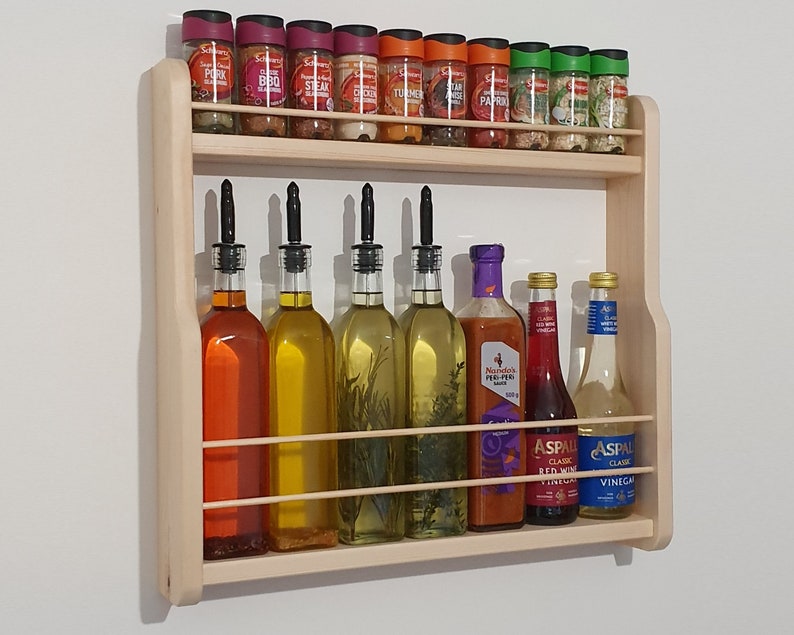 May include: A wooden wall-mounted spice rack with two shelves. The top shelf holds various spice jars with labels like "Pork Steak", "Chicken", "Turmeric", "Paprika", and "Garlic". The bottom shelf holds bottles of oil and vinegar.