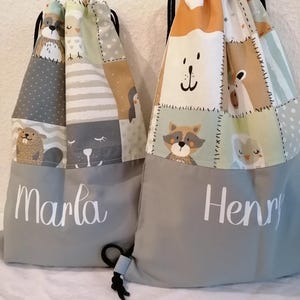 Könnte beinhalten: Zwei graue Kordelbeutel mit bunten Patchwork-Designs mit Tierillustrationen. Die Beutel haben einen grauen Bodenbereich mit den Namen "Marla" und "Henry" in weiß.