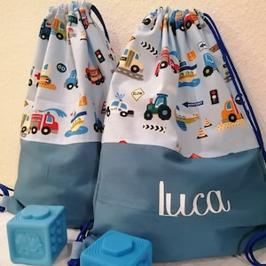 Könnte beinhalten: Zwei blaue Kordelzugbeutel mit einem hellblauen Oberteil mit farbenfrohen Illustrationen von Fahrzeugen. Die Beutel haben eine einfarbige blaue Basis und blaue Kordelzüge. Der Name "Luca" ist weiß auf einer Tasche aufgedruckt. Zwei blaue Spielzeugblöcke befinden sich im Vordergrund.