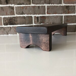 May include: A dark brown wooden riser with a flat top and curved legs. The riser is made of solid wood and has a rustic finish.