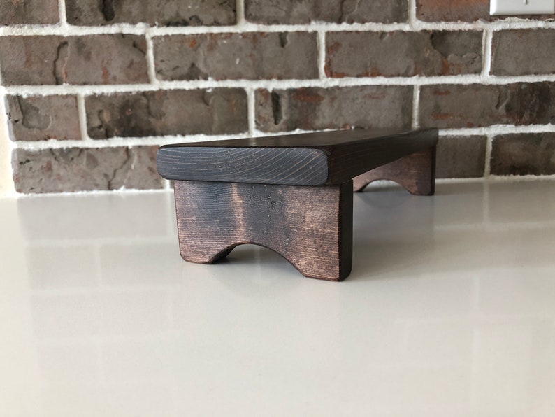 May include: A dark brown wooden riser with a flat top and curved legs. The riser is made of solid wood and has a rustic finish.