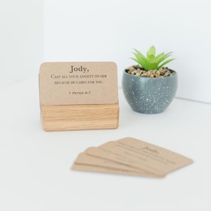 May include: A set of brown cards with the text "Jody, Cast all your anxiety on him because he cares for you. 1 Peter 5:7" on the top card. The cards are stacked on a wooden block and a small potted succulent plant is in the background.