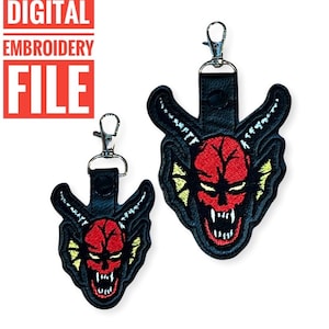 May include: Two embroidered keychains featuring a red devil face with yellow and white accents. The keychains are black and have a silver metal clasp.