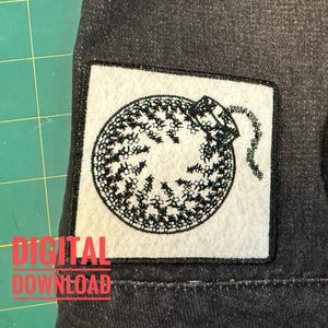 Puede incluir: Un parche cuadrado de tela blanca con un diseño bordado en negro de una bomba redonda con una mecha encendida. El parche está cosido sobre una tela de mezclilla gris oscuro. El diseño está creado con un estilo de punto de cruz, mostrando una forma circular con una pequeña mecha que sobresale por el lado superior derecho.