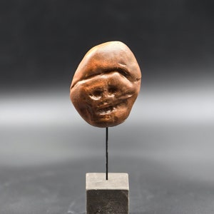 Puede incluir: Una pequeña escultura de cerámica marrón de una cara con un diseño simple y tallado. La escultura está montada sobre una pequeña base cuadrada negra.