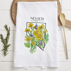 May include: White kitchen towel featuring a watercolor illustration of Nevada wildflowers. The design includes yellow and purple flowers within a black outline of the state, with the text "NEVADA wildflowers" above.