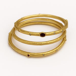 Op de afbeelding: Een set van drie gestapelde gouden armbanden met een matte, gehamerde textuur. De armbanden zijn versierd met kleine, ronde, heldere stenen en twee ovale, donkerrode edelstenen die met tussenpozen op de cirkelvormige banden zijn geplaatst.