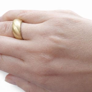 May include: A gold-colored ring with a rounded band is worn on a finger. The ring has a matte finish and a textured edge. The hand is in a relaxed position, showcasing the ring's design. The ring appears to be a wedding band or a similar type of jewelry.