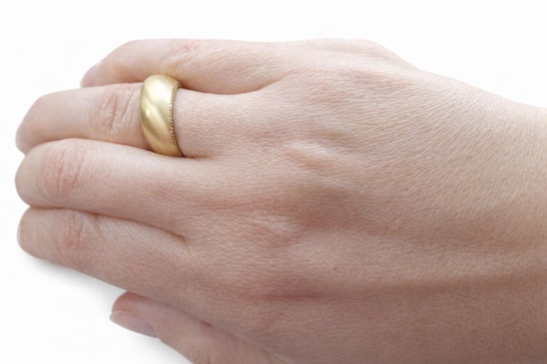 May include: A gold-colored ring with a rounded band is worn on a finger. The ring has a matte finish and a textured edge. The hand is in a relaxed position, showcasing the ring's design. The ring appears to be a wedding band or a similar type of jewelry.