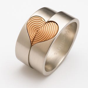 May include: Two-piece silver rings with a heart design. The heart is formed by the two rings coming together, with a textured, golden-brown heart shape in the center. The rings have a smooth, matte finish.