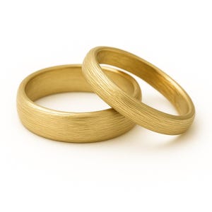 May include: Two gold wedding bands with a textured, brushed finish. One ring is positioned in front of the other, slightly angled. The rings have a rounded shape and a warm, golden hue, set against a white background.