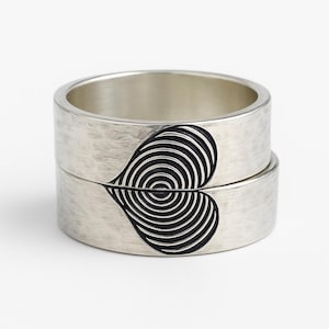 May include: Two silver rings stacked together, each with a textured surface. One side of each ring features a black heart design with concentric circles. The rings are displayed against a white background.